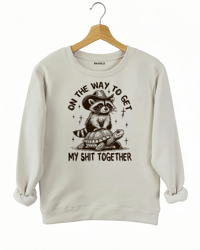 Playful raccoon and turtle design with the text 'On the Way to Get My Shit Together' on a beige sweatshirt, available on Ragglo.