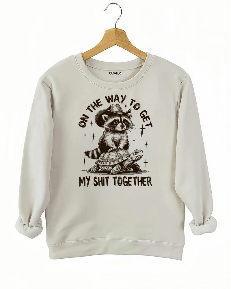 Playful raccoon and turtle design with the text 'On the Way to Get My Shit Together' on a beige sweatshirt, available on Ragglo.