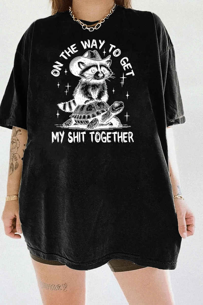 Funny graphic of a raccoon on a turtle with the text 'On the Way to Get My Shit Together' on a black T-shirt, available on Ragglo.