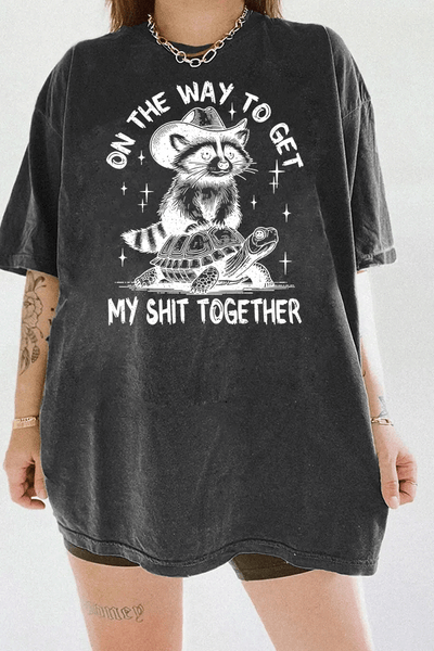 Funny graphic of a raccoon on a turtle with the text 'On the Way to Get My Shit Together' on a dark gray T-shirt, available on Ragglo.