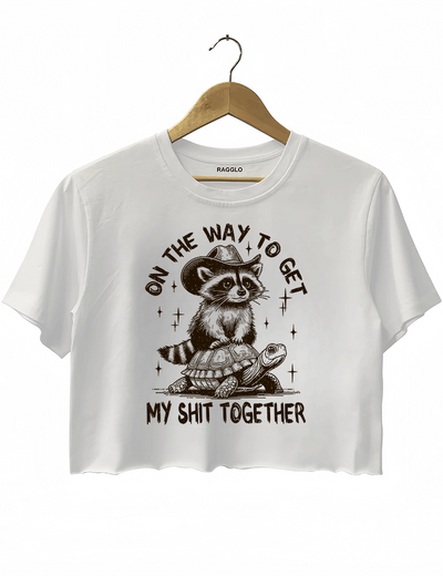 On the Way to Get My Shit Together design showing a cowboy raccoon on a turtle with playful lettering on a white crop top, available on Ragglo.