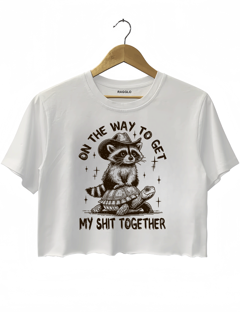 On the Way to Get My Shit Together design showing a cowboy raccoon on a turtle with playful lettering on a white crop top, available on Ragglo.