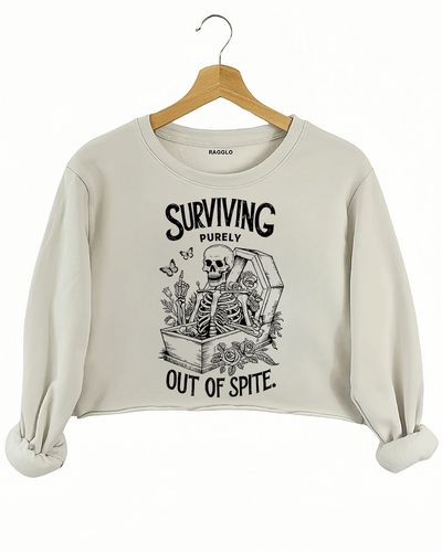 Beige crop sweatshirt with a graphic of a skeleton sitting in a coffin, adorned with roses and candles, featuring the phrase 'Surviving Purely Out of Spite,' available on Ragglo.
