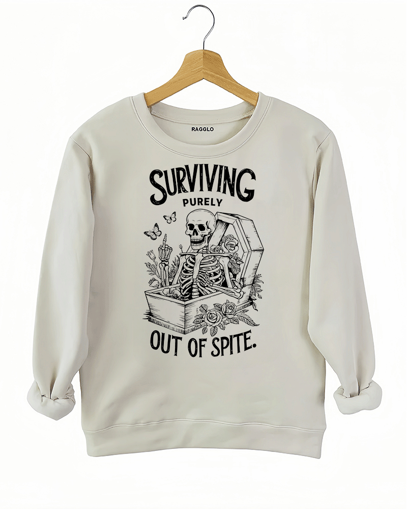 Beige sweatshirt featuring a skeleton reclining in a coffin with floral accents, butterflies, and candles, accompanied by the humorous text 'Surviving Purely Out of Spite,' available on Ragglo.