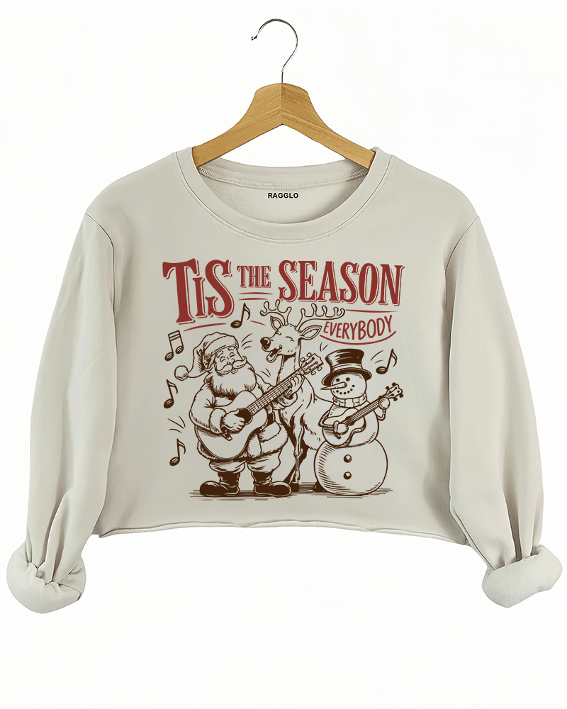 Christmas graphic with the text Tis The Season Everybody, featuring Santa, a reindeer, and a snowman playing guitars, surrounded by musical notes, printed on a beige crop sweatshirt available at Ragglo.