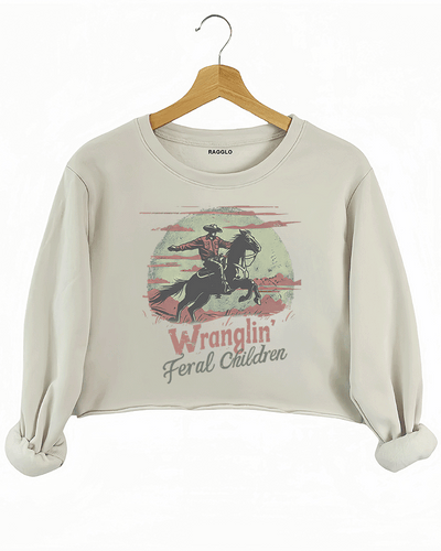 Ragglo beige crop sweatshirt displaying a retro-inspired cowboy graphic with the text 'Wranglin' Feral Children,' featuring a cowboy in a desert landscape with mountains and a textured moon.